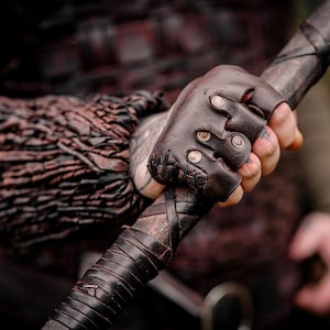 May include: A dark brown leather fingerless glove featuring metallic studs and stitched detailing, worn on a hand gripping a dark, leather-wrapped staff. The glove is paired with a textured, layered dark brown leather forearm bracer.