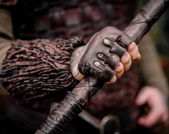 May include: A dark brown leather fingerless glove featuring metallic studs and stitched detailing, worn on a hand gripping a dark, leather-wrapped staff. The glove is paired with a textured, layered dark brown leather forearm bracer.