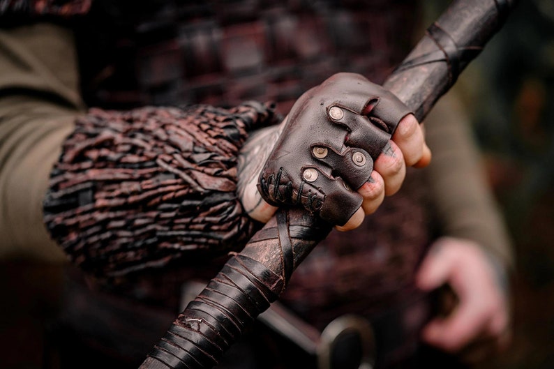 May include: A dark brown leather fingerless glove featuring metallic studs and stitched detailing, worn on a hand gripping a dark, leather-wrapped staff. The glove is paired with a textured, layered dark brown leather forearm bracer.