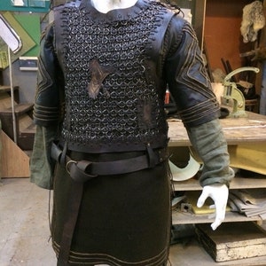 May include: A dark brown leather and metal chainmail tunic featuring a central raven emblem on the chest. The garment includes short sleeves with stitched detailing, a matching leather belt with a metal ring buckle, and a long dark tunic base.