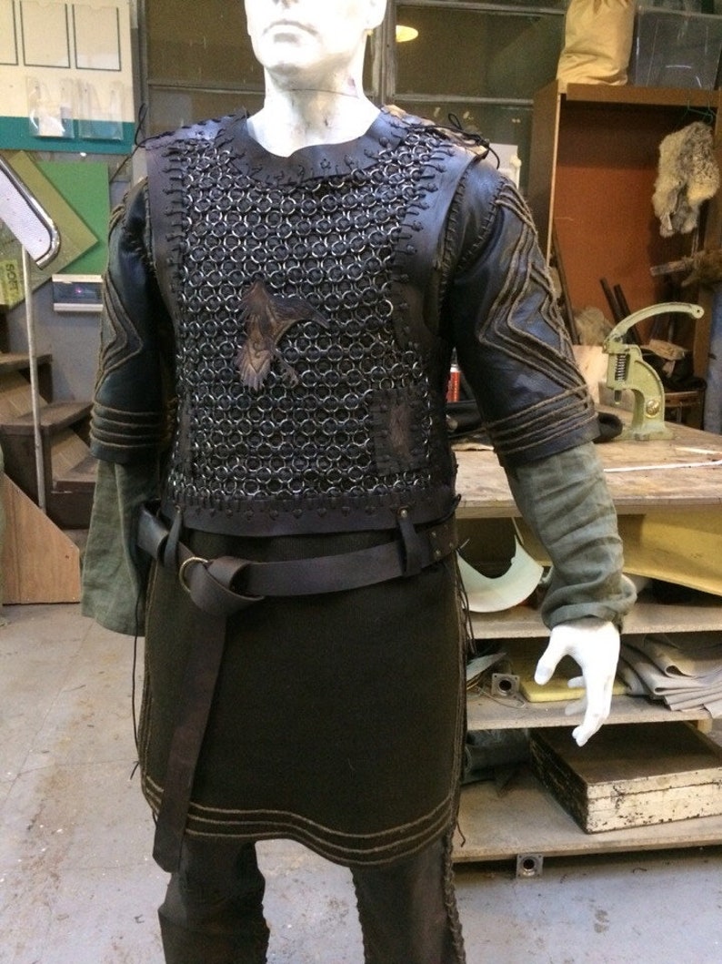 May include: A dark brown leather and metal chainmail tunic featuring a central raven emblem on the chest. The garment includes short sleeves with stitched detailing, a matching leather belt with a metal ring buckle, and a long dark tunic base.