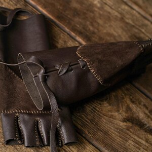 May include: A pair of dark brown fingerless leather gloves featuring a suede panel on the back of the hand, visible stitching along the edges, and a leather lace-up closure system on the wrist area to adjust the fit securely around the forearm.