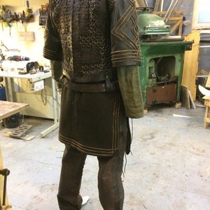 May include: A dark brown leather and metal chainmail vest worn over a long-sleeved olive green tunic. The outfit includes a dark brown leather belt, matching trousers with side lacing, and leather shoulder pieces featuring stitched geometric patterns.