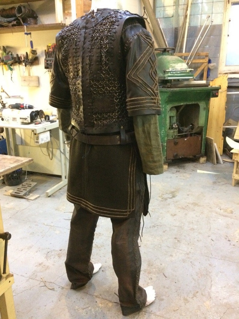 May include: A dark brown leather and metal chainmail vest worn over a long-sleeved olive green tunic. The outfit includes a dark brown leather belt, matching trousers with side lacing, and leather shoulder pieces featuring stitched geometric patterns.