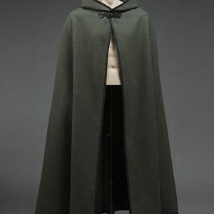 May include: A floor-length, dark olive green wool-textured cloak featuring a hood, black trim along the edges, and a single black leather-style buckle closure at the neck. The garment is displayed on a mannequin, showing a full-length silhouette.