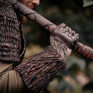 May include: A dark brown leather arm guard and shoulder piece featuring a woven, textured design. The arm guard is worn on the forearm, paired with a fingerless leather glove. A wooden axe handle with leather wrapping is held in the hand.