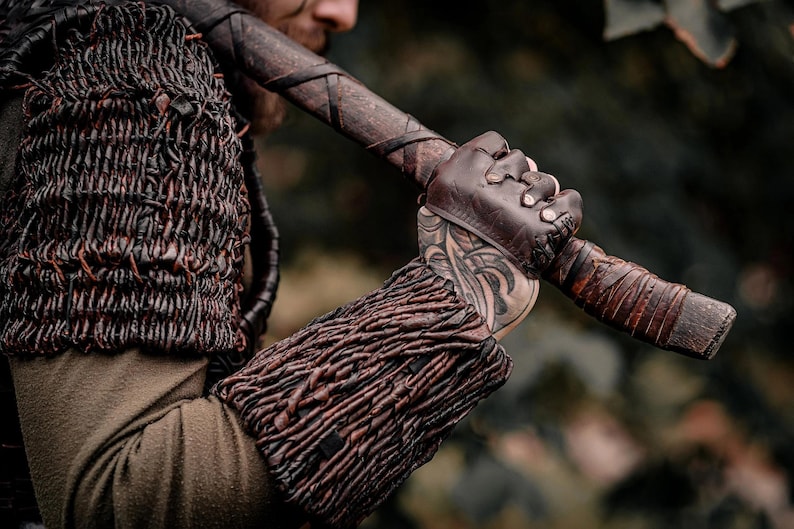 May include: A dark brown leather arm guard and shoulder piece featuring a woven, textured design. The arm guard is worn on the forearm, paired with a fingerless leather glove. A wooden axe handle with leather wrapping is held in the hand.
