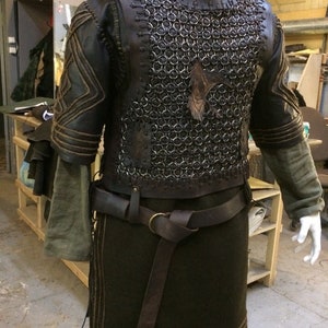 May include: A dark brown leather and metal chainmail vest worn over a long-sleeved olive green tunic. The vest features leather lacing, a dark leather belt with a brass ring buckle, and a leather raven emblem attached to the chest area.