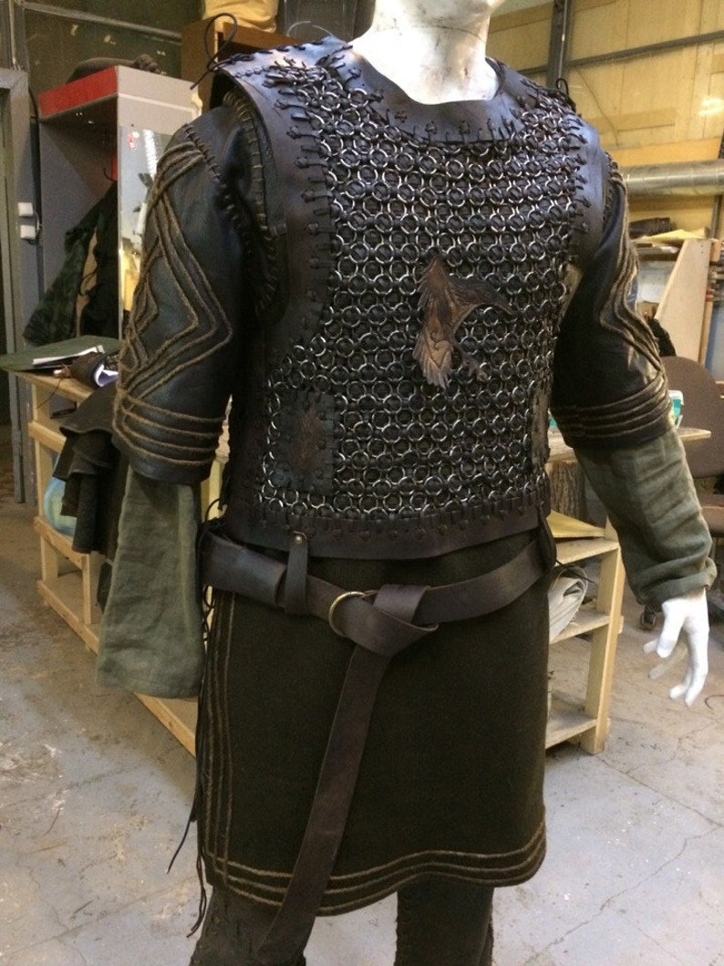 May include: A dark brown leather and metal chainmail vest worn over a long-sleeved olive green tunic. The vest features leather lacing, a dark leather belt with a brass ring buckle, and a leather raven emblem attached to the chest area.