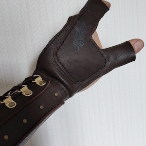 May include: A dark brown leather fingerless glove featuring a reinforced palm patch with a subtle embossed feather design. The wrist section includes a lace-up closure with brass-toned metal eyelets and a series of small brass-toned rivets along the side.