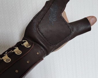May include: A dark brown leather fingerless glove featuring a reinforced palm patch with a subtle embossed feather design. The wrist section includes a lace-up closure with brass-toned metal eyelets and a series of small brass-toned rivets along the side.