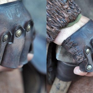 May include: A pair of fingerless dark brown leather gloves featuring a distressed finish, decorative cross-stitching along the side, and metallic brass-toned studs securing the individual finger sections across the knuckles for a structured fit.