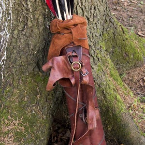 May include: A brown leather quiver containing several wooden arrows with black and red fletching. The quiver features a soft suede top section, adjustable leather straps with brass-colored metal buckles, and a crisscross lacing design along the body.