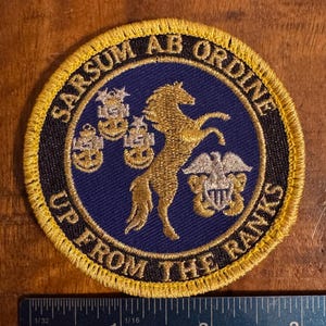 May include: A gold embroidered patch with a black background and a gold border. The patch features a gold horse rearing up on its hind legs with three gold anchors and a gold eagle with a silver shield. The text "Sarsum Ab Ordine Up From The Ranks" is embroidered in gold around the edge of the patch.