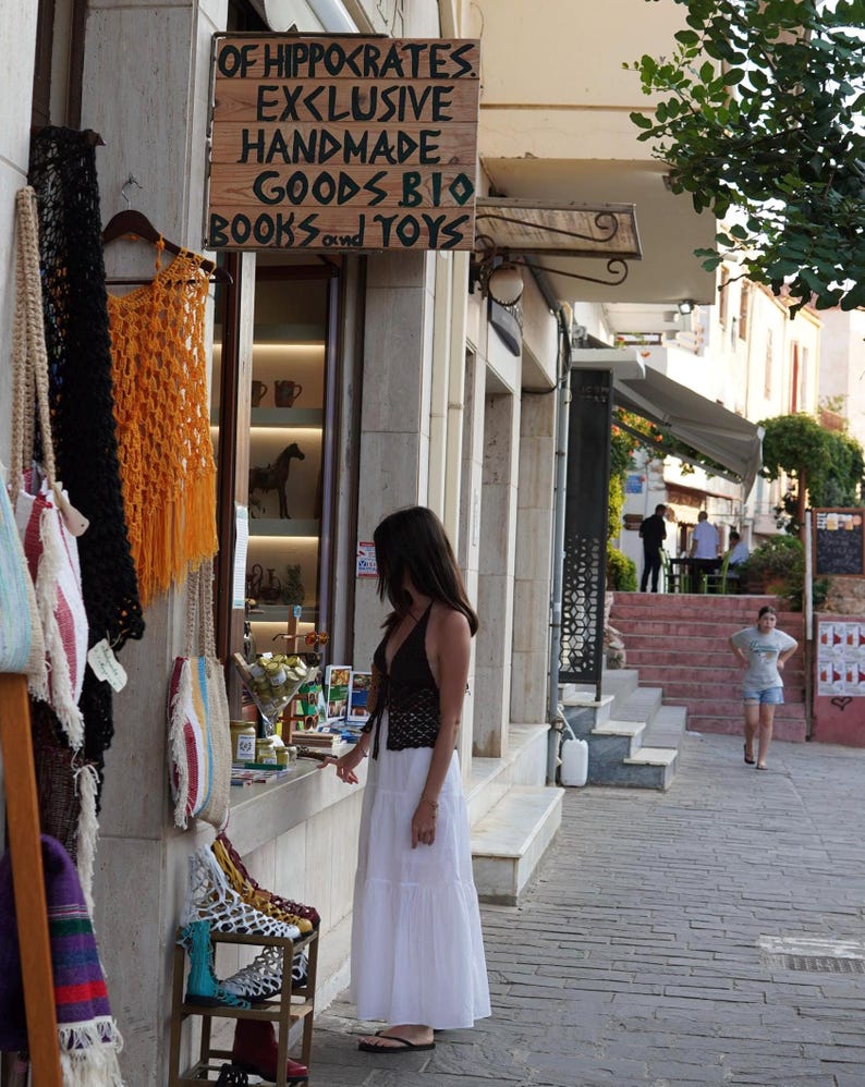 Pu&ograve; includere: Scena di strada con un negozio con un'insegna di legno che recita "OF HIPPOCRATES EXCLUSIVE HANDMADE GOODS BIO BOOKS and TOYS". Una donna con un vestito bianco e un top nero &egrave; di fronte al negozio. Sono esposti vari oggetti fatti a mano.