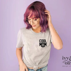 Op de afbeelding: Een grijs T-shirt met de tekst "COOL MOM" in zwart gedrukt op de voorkant. Het shirt wordt gedragen door een persoon met roze haar en een bril.