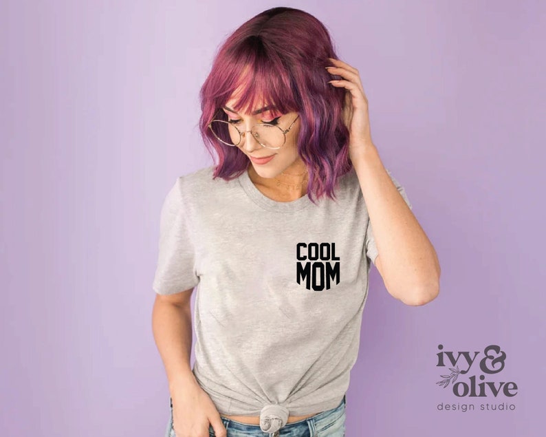 Op de afbeelding: Een grijs T-shirt met de tekst "COOL MOM" in zwart gedrukt op de voorkant. Het shirt wordt gedragen door een persoon met roze haar en een bril.