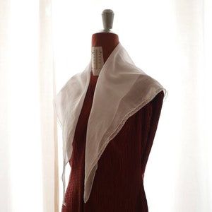 Puede incluir: Una bufanda blanca de gasa con ribete de encaje drapeada sobre un maniquí de vestido rojo.