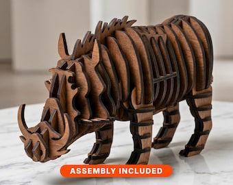 May include: A brown wooden 3D puzzle sculpture of a warthog, constructed from multiple interlocking laser-cut panels. The animal figure features prominent tusks and a layered, ribbed design. An orange rectangular label reads ASSEMBLY INCLUDED.