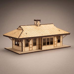 Pode incluir: Uma maquete em miniatura de uma estação ferroviária construída em madeira de cor clara. A estrutura apresenta um telhado de telhas, uma chaminé central, várias janelas com padrão de grade e beirais salientes apoiados por suportes de madeira.