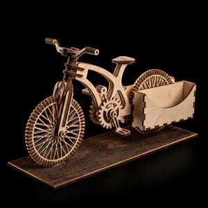 May include: A laser-cut wooden bicycle model featuring a light-colored frame, spoked wheels, and a gear mechanism. The bicycle is attached to a rectangular wooden base and includes a small, open-topped storage box mounted over the rear wheel.