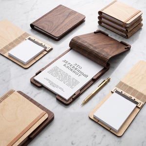 Può includere: Una collezione di taccuini e blocchi per appunti in legno con copertine in noce scuro e acero chiaro. Un taccuino aperto mostra carta bianca con il testo "ЭТО ДЕРЕВЯННЫЙ БЛОКНОТ" e una penna in metallo dorato appoggiata accanto.