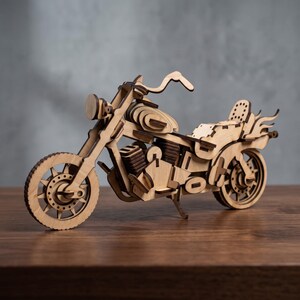 Puede incluir: Una maqueta de motocicleta de madera clara con un marco detallado, ruedas de radios, depósito de combustible, bloque motor, asiento con respaldo y detalles en forma de llama en el guardabarros trasero, hecha de piezas de madera cortadas a láser.