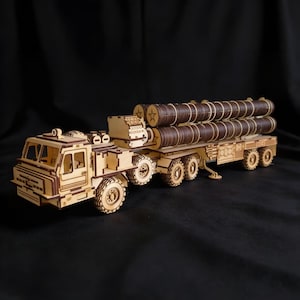 Pode incluir: Um modelo de madeira cortado a laser de um caminhão de transporte militar com um reboque de vários eixos transportando vários componentes cilíndricos em forma de míssil. O modelo exibe uma construção complexa em camadas.