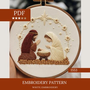Può includere: Scena della natività ricamata in un cerchio di legno. Il disegno presenta Maria, Giuseppe e Gesù bambino in una mangiatoia, circondati da una ghirlanda floreale. Il testo "EMBROIDERY PATTERN" e "WHITE.EMBROIDERY" sono visibili.