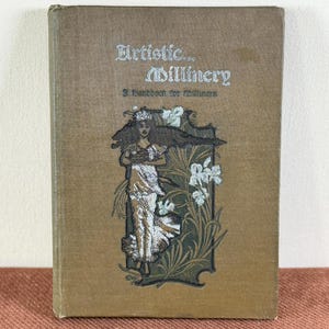 May include: A hardcover book titled Artistic Millinery: A Handbook for Milliners with a brown fabric cover. The front cover features a silver-toned illustration of a person wearing a gown surrounded by stylized floral elements and foliage.