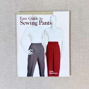 May include: A book titled "Easy Guide to Sewing Pants" from the Sewing Companion Library. The cover features two mannequins, one wearing gray pants and the other wearing red pants. The author's name, Lynn MacIntyre, is also visible.