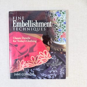 May include: A book titled "FINE Embellishment TECHNIQUES" with the subtitle "Classic Details for Today's Clothing" by Jane Conlon. The cover features a red garment with cream-colored embroidery and a decorative vase.