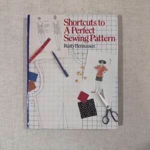May include: A sewing book titled "Shortcuts to A Perfect Sewing Pattern" by Rusty Bensussen. The cover features illustrations of clothing, sewing patterns, and sewing tools, including scissors, a pen, and fabric swatches. The book is on a grid background.