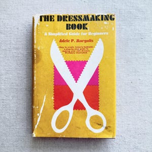 May include: A vintage book titled "The Dressmaking Book" with a yellow cover. The cover features white scissors over a pink and red fabric design. Text includes "A Simplified Guide for Beginners" and author Adele P. Margolis.