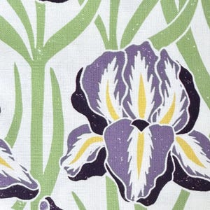 May include: Close-up of a fabric with a floral pattern. The design features a large purple iris flower with yellow accents and dark purple outlines. Green, stylized leaves are in the background, all set against a white backdrop.