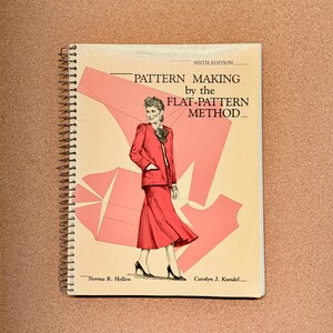 May include: A spiral-bound book titled "PATTERN MAKING by the FLAT-PATTERN METHOD." The cover features a red illustration of a woman in a red suit and skirt. The book is a sixth edition, with the authors' names at the bottom.