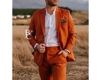 May include: A burnt orange suit with a white dress shirt and a patterned pocket square. The suit jacket has a two-button closure and a notched lapel. The person is standing in a field of tall grass.