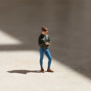 May include: A miniature figurine of a person wearing a dark green jacket, blue jeans, and brown shoes. The person is holding a small object in their hand.
