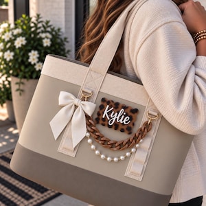 May include: A beige and dark taupe tote bag featuring a cream-colored fabric top panel and matching shoulder straps. The bag is decorated with a white satin bow, a brown chain link accent, a string of white pearls, and a leopard print name tag.