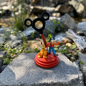 May include: A red and black Mickey Mouse-shaped figurine with a yellow and brown Goofy figurine standing on top of it. The figurines are on a red base.