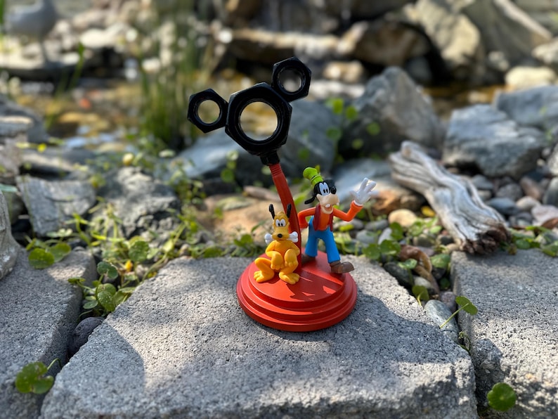 May include: A red and black Mickey Mouse-shaped figurine with a yellow and brown Goofy figurine standing on top of it. The figurines are on a red base.
