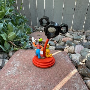 May include: A red and black Mickey Mouse themed phone stand with a black metal base and a red plastic base. The stand features a Mickey Mouse head made of nuts and bolts. A small plastic figurine of Mickey Mouse and Pluto is attached to the base.