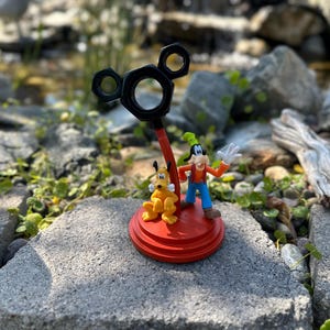 May include: A red and black Mickey Mouse-shaped stand with a yellow and orange Goofy figurine and a yellow and orange Pluto figurine. The stand is on a gray stone surface.