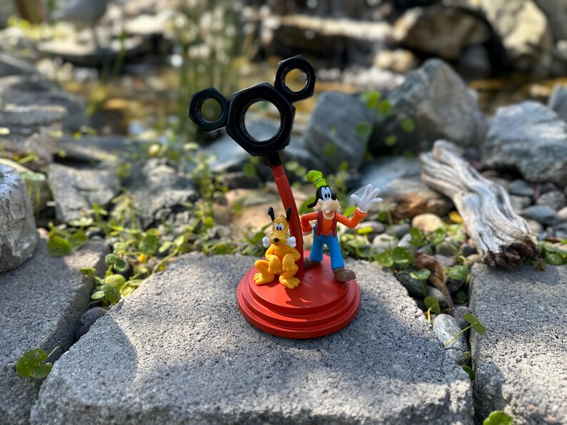 May include: A red and black Mickey Mouse-shaped stand with a yellow and orange Goofy figurine and a yellow and orange Pluto figurine. The stand is on a gray stone surface.