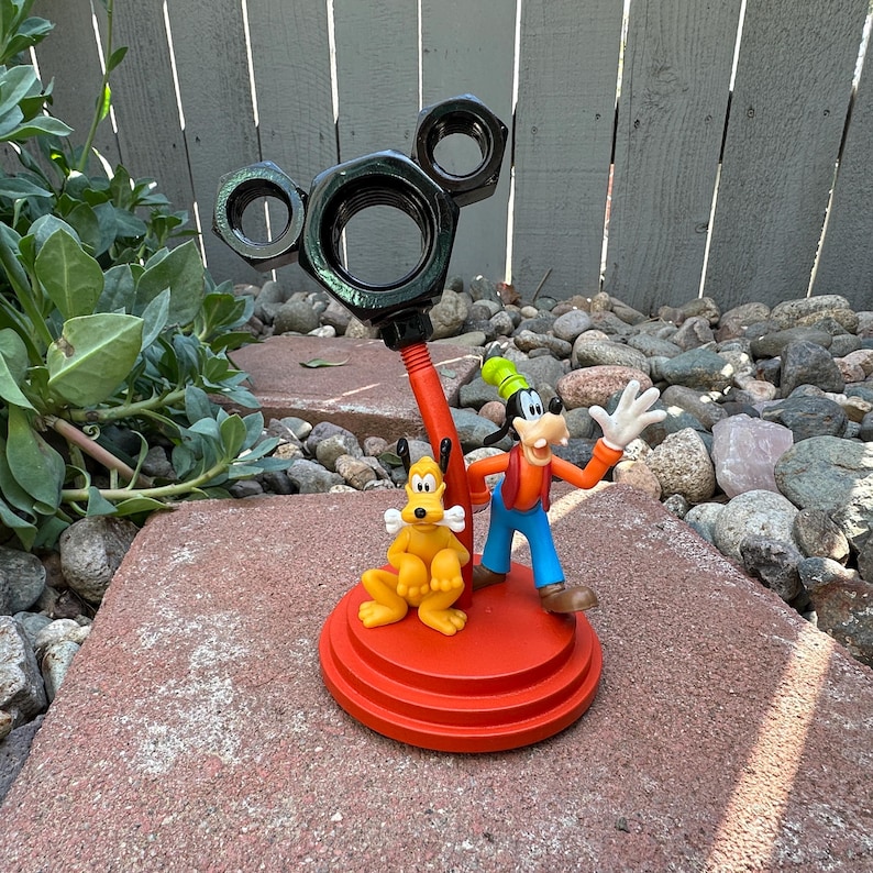 May include: A red and black metal Mickey Mouse-shaped stand with a Goofy and Pluto figurine on top. The stand is made of nuts and bolts and has a red base.