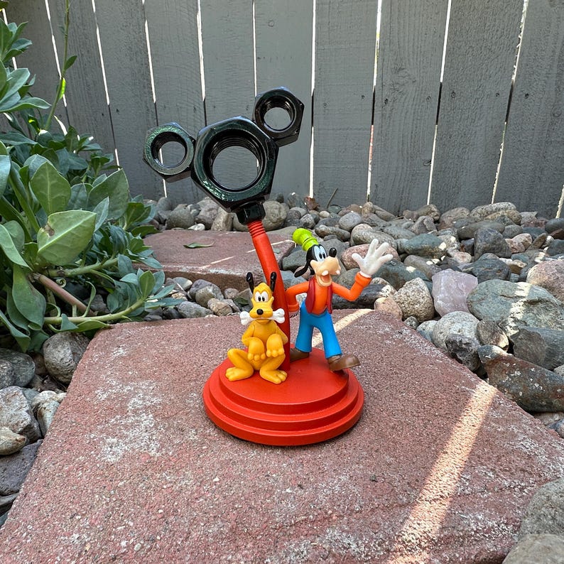 May include: A red and black metal Mickey Mouse-shaped stand with a red base.  Goofy and Pluto figurines are standing on the base.  Goofy is wearing a red and blue outfit and a green hat.  Pluto is wearing a yellow and orange outfit.