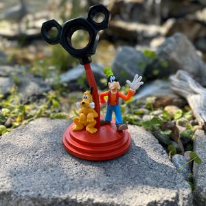 May include: A red and black Mickey Mouse themed figurine featuring Goofy and Pluto. Goofy is wearing a red and blue outfit and a green hat. Pluto is a yellow dog with a black nose and tail. The figurine is made of plastic and is about 6 inches tall.