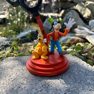 May include: A red and black desk organizer featuring a cartoon Goofy and Pluto figurine. Goofy is waving and Pluto is holding a bone. The organizer is on a gray stone surface.