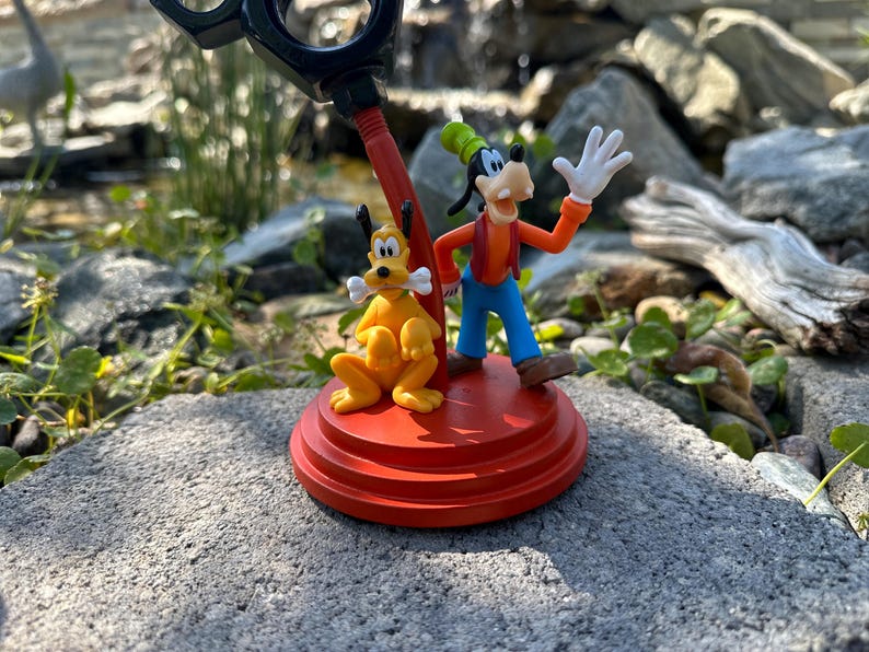 May include: A red and black desk organizer featuring a cartoon Goofy and Pluto figurine. Goofy is waving and Pluto is holding a bone. The organizer is on a gray stone surface.