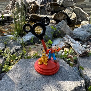 May include: A red and black Mickey Mouse-shaped desk organizer with a Goofy and Pluto figurine. The organizer has a base with three tiers and a Mickey Mouse head with ears on top. The figurine is of Goofy and Pluto standing on the base.